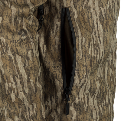 Close-up of MST Windproof Softshell Jacket with Mossy Oak Bottomland camo, showcasing a partially unzipped pocket and logo, highlighting its hunting functionality.