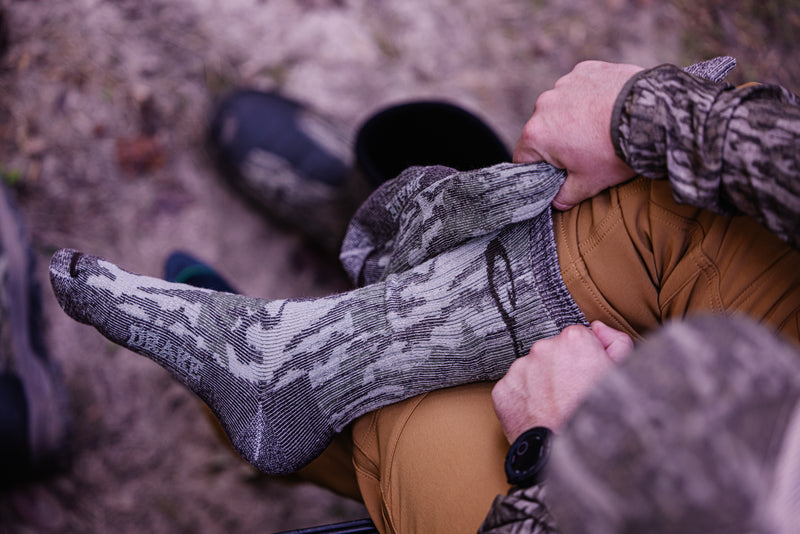 A person outdoors pulls up a Midweight Merino Camo Mid Calf Hunting Sock over their foot, showcasing its camo pattern and durable design.