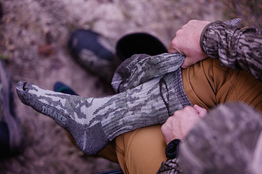 A person outdoors pulls up a Midweight Merino Camo Mid Calf Hunting Sock over their foot, showcasing its camo pattern and durable design.