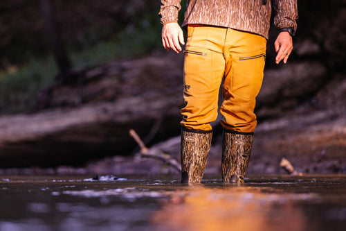 Person waist-deep in water wearing MST CommandXT Softshell Pant with waterproof features and camouflage wading boots, showcasing mobility and protection for demanding outdoor activities.