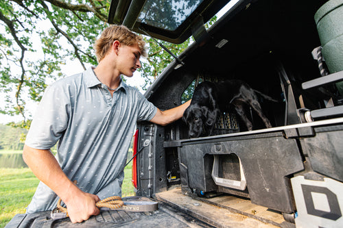 Young man in a Performance Print Polo Shirt guides a puppy from a truck's DECKED storage, implying outdoor adventure readiness.