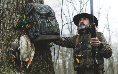 Outdoorsman in camouflage gear wearing the MST CommandXT Softshell Vest, holding a shotgun, with a duck string and pack on a winter woodland tree.