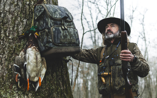 Outdoorsman in camouflage gear wearing the MST CommandXT Softshell Vest, holding a shotgun, with a duck string and pack on a winter woodland tree.