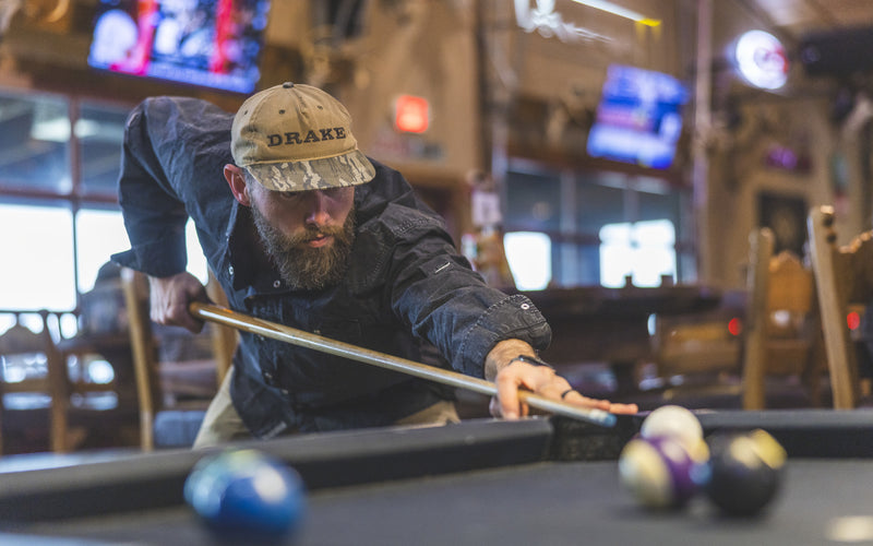 A man in a casual setting lines up a pool shot, wearing the Drake Cinco Ranch Western Solid Acid Wash Long Sleeve Shirt, showcasing its stylish, comfortable design.