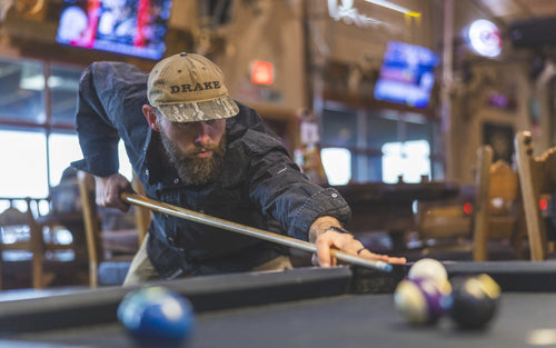 A man in a casual setting lines up a pool shot, wearing the Drake Cinco Ranch Western Solid Acid Wash Long Sleeve Shirt, showcasing its stylish, comfortable design.