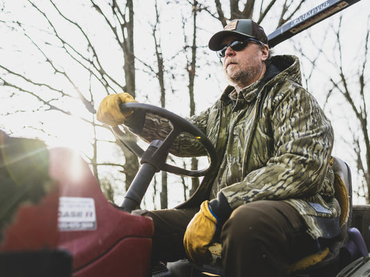 A man wearing an insulated, camouflage hooded Duck Canvas Work Jacket and yellow gloves drives a tractor.