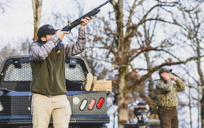 Man wearing Camp Fleece Vest aims shotgun, standing near utility vehicle with shooting gear; highlights vest's warmth, durability, and practical design for outdoor sports.