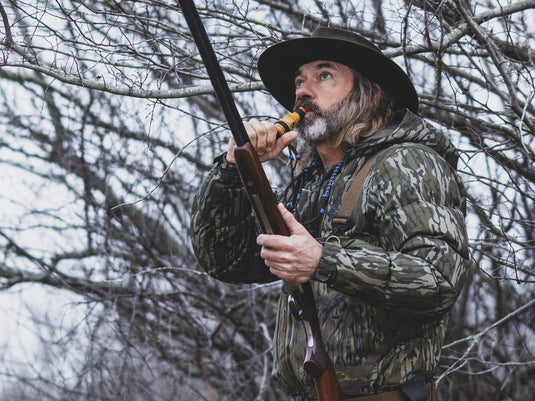 Hunter in a leafless woodland wears Drake BMF Down Jacket, using a game call with a shotgun in hand, showcasing hunting gear durability and warmth.