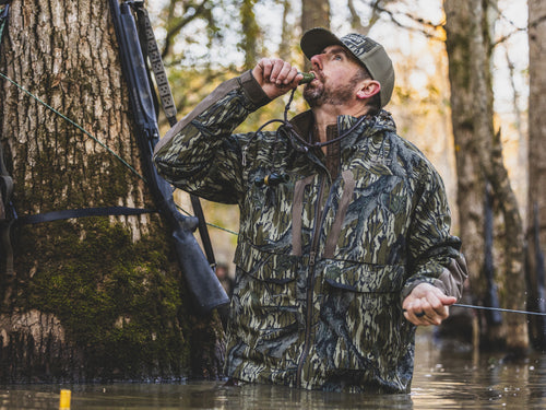 Outdoorsman in swamp wearing LST BMF Pit Jacket, using a duck call, showcasing its durability and warmth; shotgun visible nearby.