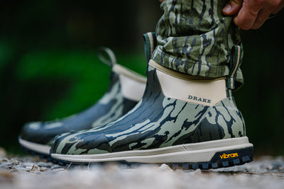 Drake Offroad Rubber Deck Boots with camouflage pattern, featuring Vibram soles and neoprene lining, shown outdoors with a hand adjusting camo pants.