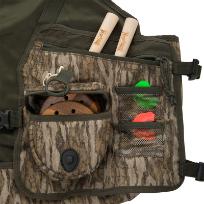 Turkey Strap Vest front with camo pattern, displaying maple-wood game calls, fly-fishing reel, and mesh pockets with green and orange markers for hunting efficiency.