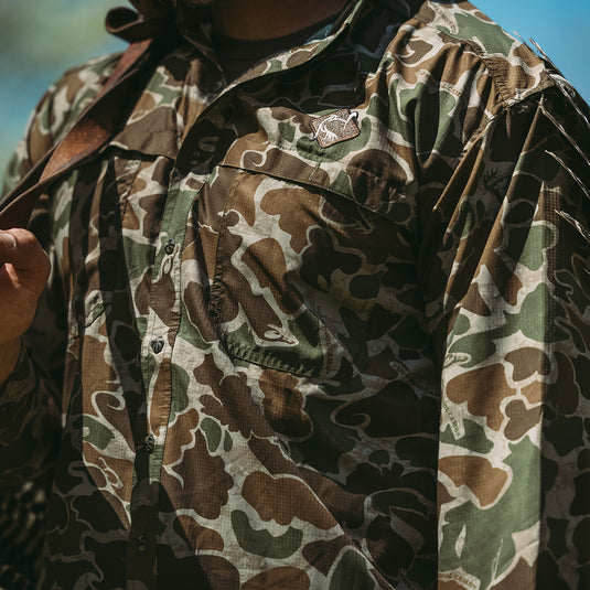 A man wearing a camouflage shirt with a bird patch and thumb close-up, showcasing the Men's Mesh Back Flyweight Turkey Shirt.