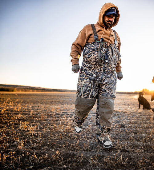 Man wearing BMF Uninsulated Pit Bib in a harvested field, showcasing durable, waterproof hunting gear, with a black dog sitting nearby at sunset.