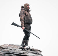 Wearing the Waterfowl LST Hardline 3-in-1 Jacket, a person in camouflage stands on rocky terrain with a rifle, three birds at their feet, set against a plain white background.