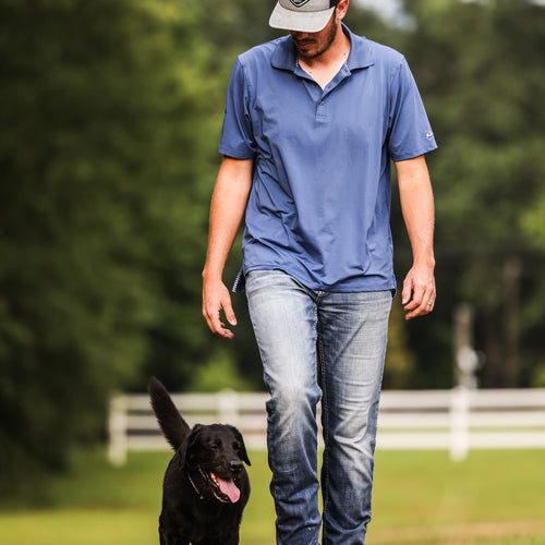 A man in a blue shirt walking a black dog outdoors. Performance Stretch Polo S/S by Drake Waterfowl. Features 4 Way Stretch, Moisture Wicking, UPF Sun Protection, and Rib Knit Collar.