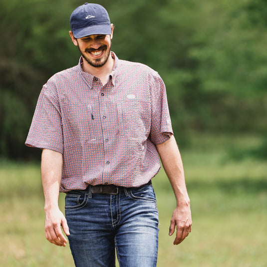 A man in a red and white checkered shirt and blue hat, showcasing the Drake Classic Seersucker Grid Check Button-Down Shirt. Features include UPF30 sun protection, moisture-wicking fabric, and hidden zippered pockets.