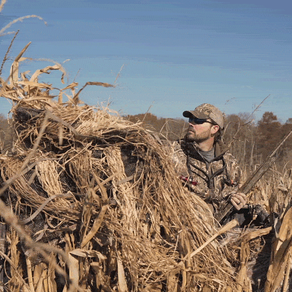 A man in a camouflaged suit holding a gun in a field, featuring the Ghillie 4-Man Blind with No-Shadow Dual Action Top.