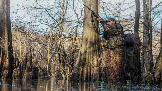 A man in camouflage holding a gun in the Ghillie Shallow Water Chair Blind, perfect for concealed duck hunting.