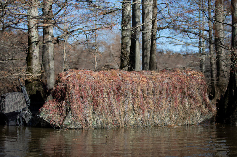 Ghillie Boat Blind with No-Shadow Dual Action Top: A large rock covered in grass in water, with trees in the background.