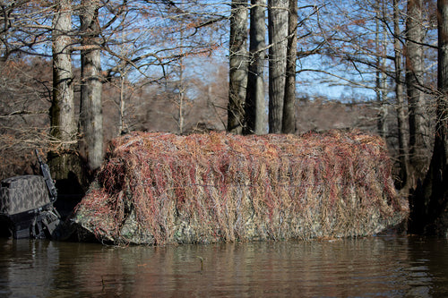 Ghillie Boat Blind with No-Shadow Dual Action Top: A large rock covered in grass in water, with trees in the background.