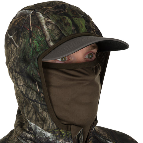 A person wearing a lightweight Pursuit Full Zip Hoodie with Agion Active XL®, featuring a camouflage pattern and a cap.