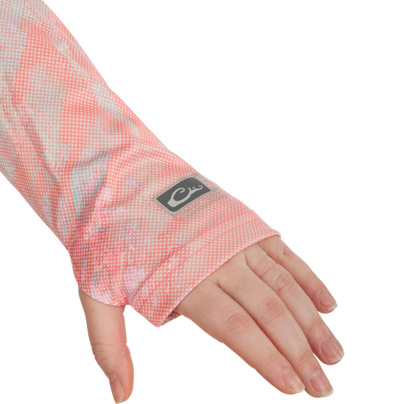 A close-up of a hand wearing a pink and white sleeve with a logo. The hand is part of the Women's Performance Crew Realtree Aspect Dot, a lightweight and breathable garment with UPF 50 sun protection, moisture-wicking, and quick-drying features. Perfect for outdoor activities like fishing and beach outings.