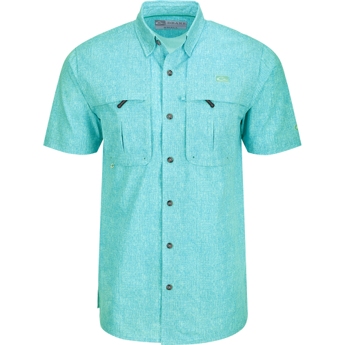 Heritage Heather Shirt S/S: A close-up of a blue shirt with pockets, featuring a logo. Lightweight, moisture-wicking, and quick-drying fabric for comfort. Hidden button-down collar and vented cape back for ventilation. Perfect for outdoor activities or office wear.