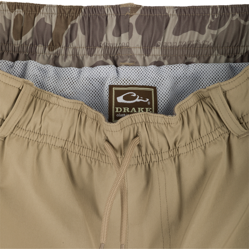 A pair of khaki Dock Shorts with a label, zipper, and fabric close-ups. Made of durable nylon/spandex blend with water-resistant finish. Features elastic waist, mesh pockets, and 6