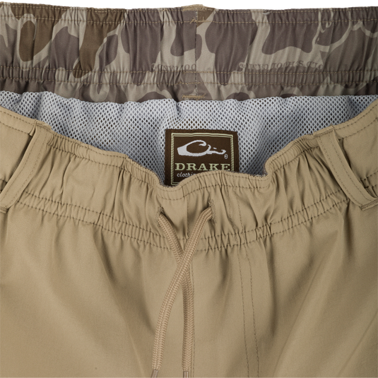 A pair of khaki Dock Shorts with a label, zipper, and fabric close-ups. Made of durable nylon/spandex blend with water-resistant finish. Features elastic waist, mesh pockets, and 6" inseam. Perfect for outdoor activities.