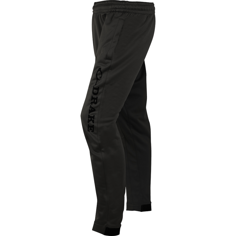 A close-up of the MST Fleece Wader Pant, a pair of black pants with a logo. Ideal for wearing under waders. Warm, comfortable, and durable. Front handwarmer pockets, elastic waist, and snug fit tapered ankles.