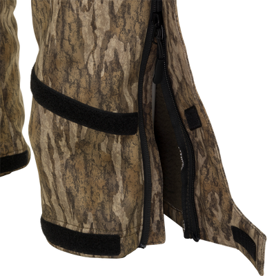 A close-up of the MST Ultimate Wader Bib, featuring a camouflage pattern, zipper, and black fabric. Sherpa-lined slash handwarmer pockets and zippered rear security pockets provide ample storage. The bib offers easy movement with its four-way stretch material, gusseted crotch, and articulated knees. Perfect for hunting and fishing.