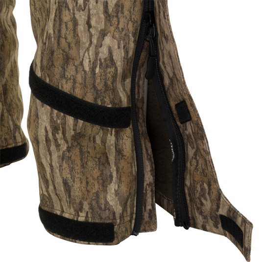 A close-up of the MST Ultimate Wader Bib, featuring a camouflage pattern, zipper, and black fabric. Sherpa-lined slash handwarmer pockets and zippered rear security pockets provide ample storage. The bib offers easy movement with its four-way stretch material, gusseted crotch, and articulated knees. Perfect for hunting and fishing.