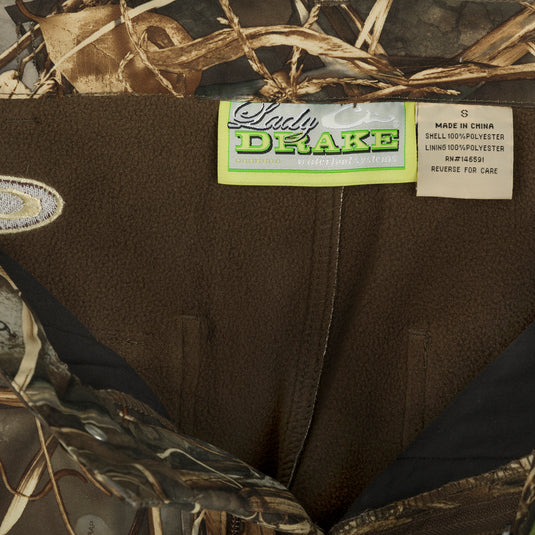 MST Women’s Refuge Bonded Fleece Pants, a close-up of camouflage pants with label, plant, and jacket details. Waterproof and windproof with fleece lining, adjustable waist, ankle cuffs, and stirrups.