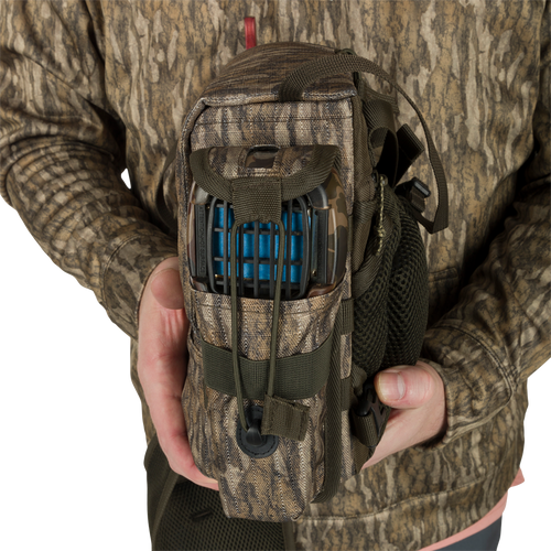 A person holding the Turkey Chest Pack, a lightweight hunting bag made with durable 600D Polyester. Features include a removable bino harness, multiple pockets and loops, and a stowable rain fly for protection. Perfect for your next hunting trip.
