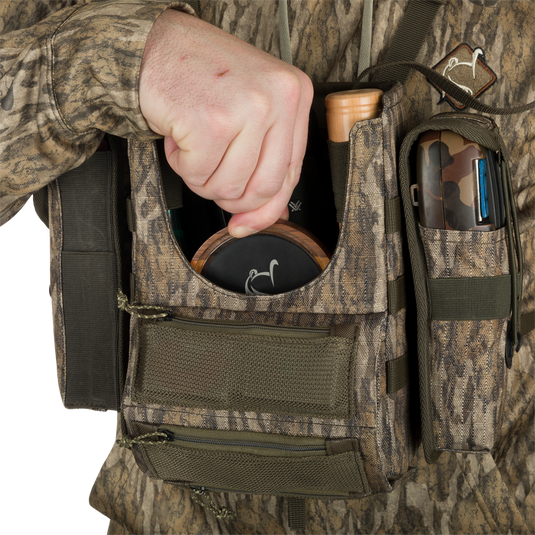 A person holding the Turkey Chest Pack, a hunting bag with multiple pockets and loops, made of durable 600D Polyester. Lightweight at 5 lbs, it won't weigh you down on your adventures. Includes a removable bino harness and a stowable rain fly for protection.